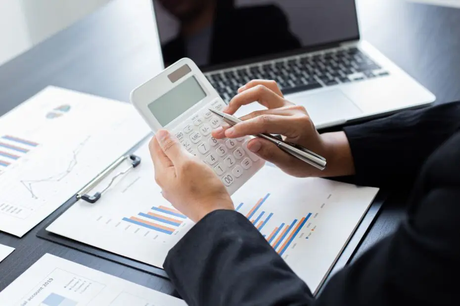 Close up of hands holding and using calculator above papers with graphs and charts and a laptop on a table