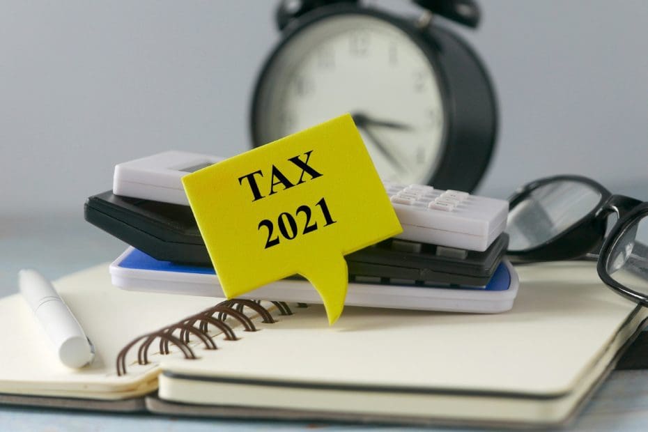 Calculators glasses and a pen sit on a notebook with Tax 2021 in front and a alarm clock in the background
