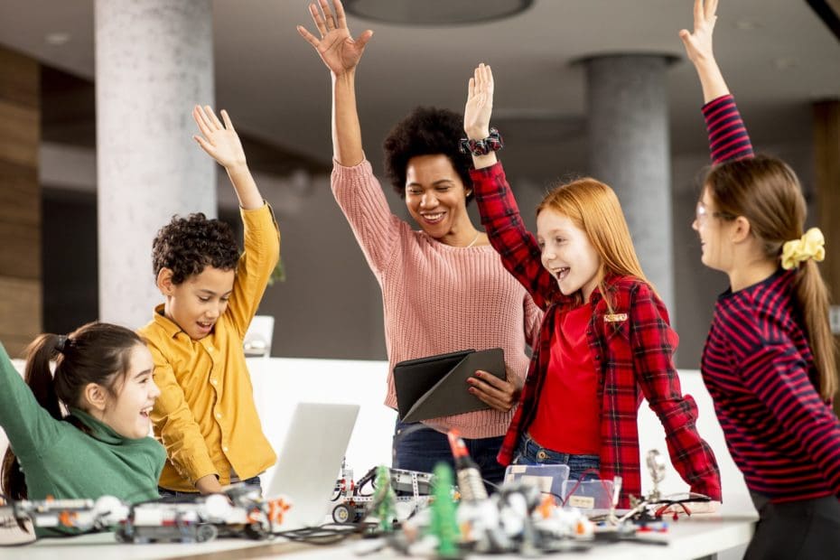 Group of happy kids with their African American female science teacher with laptop programming electric toys and robots at robotics classroom