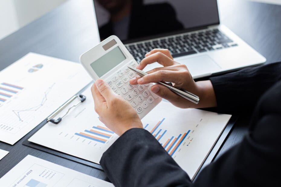 Close up of hands holding and using calculator above papers with graphs and charts and a laptop on a table